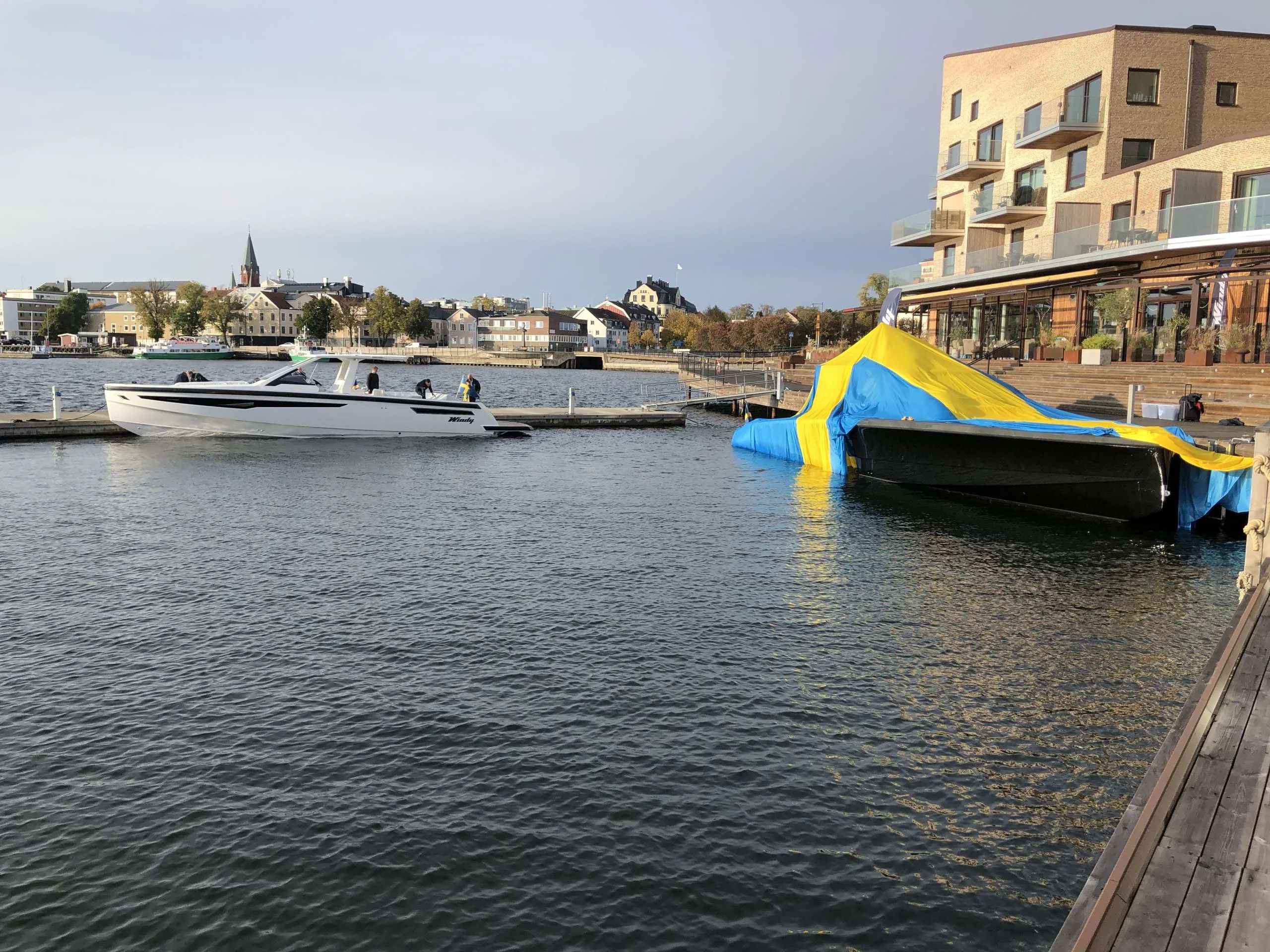 Windy SR 60 in side view, cruising in front of a pier next to a covered boat and coastal buildings.