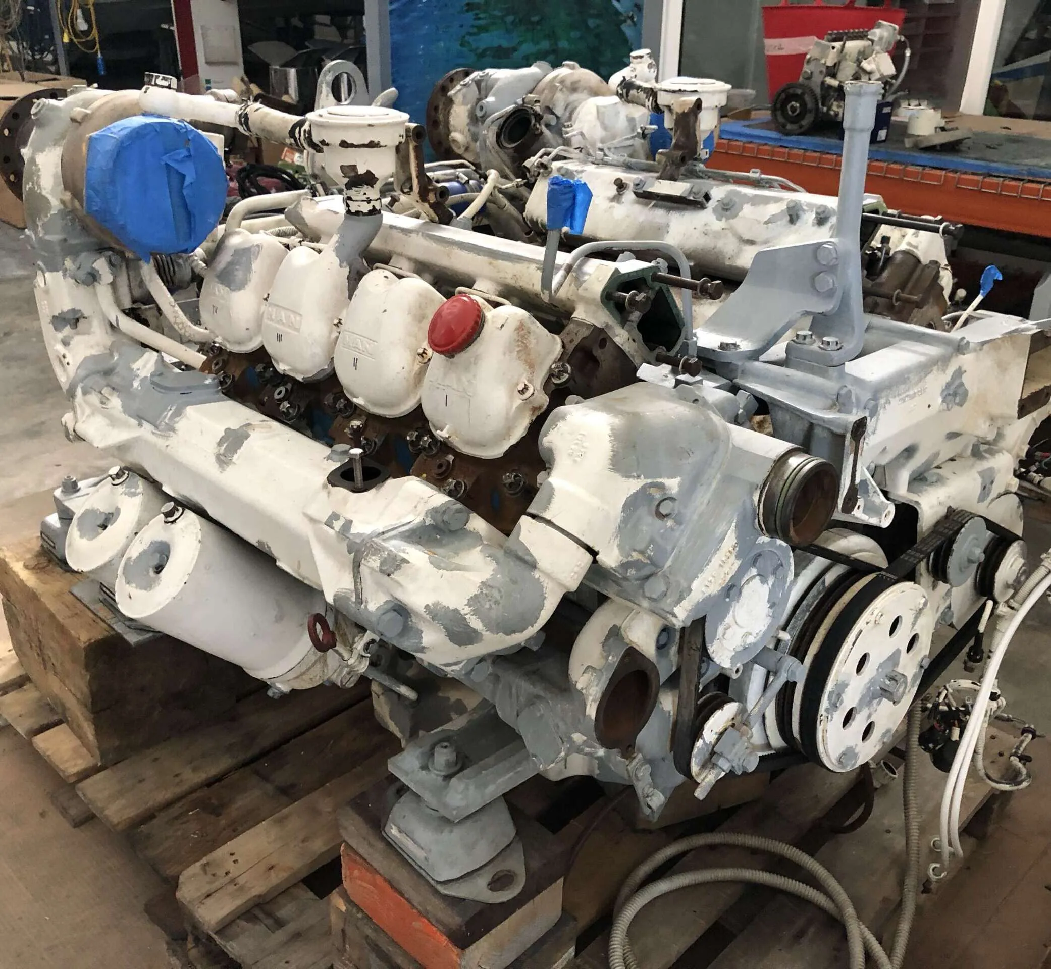 Moteur marin démonté sur un support, vu en diagonale, avec culasses, poulies, courroies et tuyaux visibles.