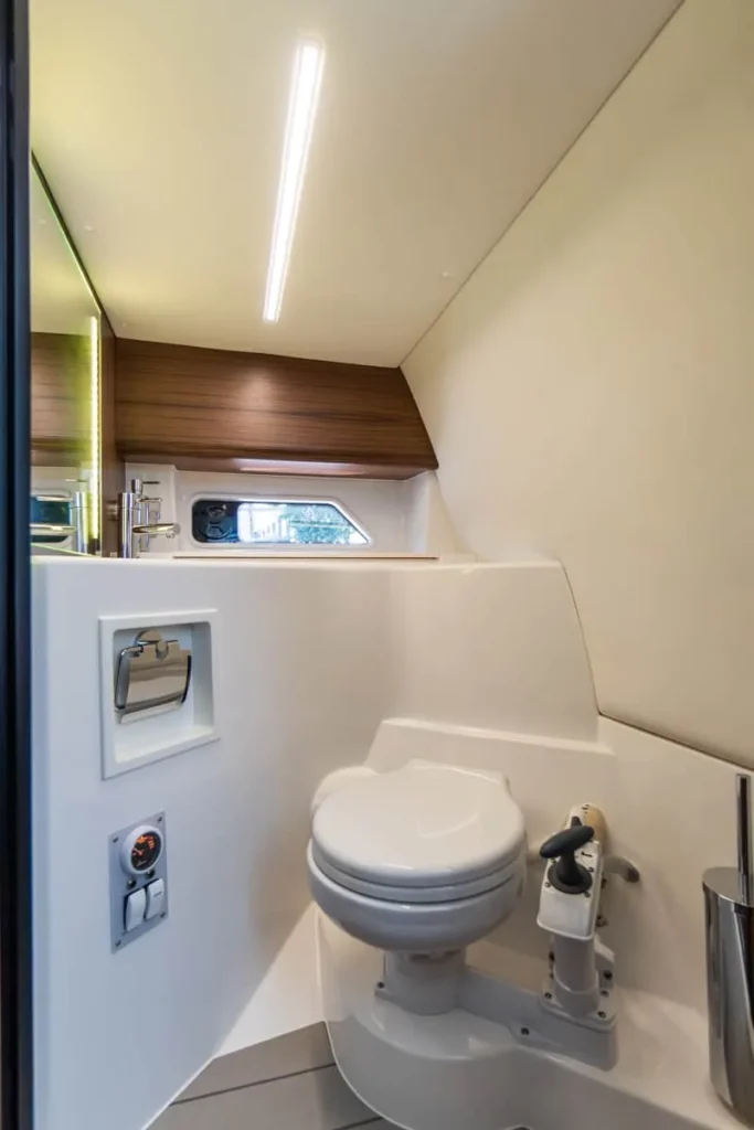 Intérieur de la salle de bain du Windy 27 Solano RS, vu de face, avec toilettes marines, fenêtre latérale et meuble en bois.