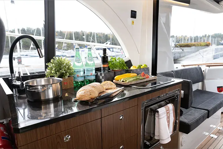 Interior de la Marex 330 Scandinavia, cocina con encimera, fregadero, fogón, alimentos y asientos junto a ventanales.