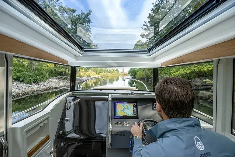 Interior del Nimbus C9, puesto de mando con pantalla náutica, patrón al timón y techo panorámico sobre un canal.