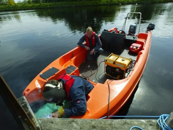 eg1 Orange Pioner Multi III viewed from above, with two people, an outboard motor and equipment on board next to a dock.