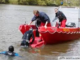 Red Pioner Multi III seen from the side, with two people, a diver, and an outboard motor on the water.
