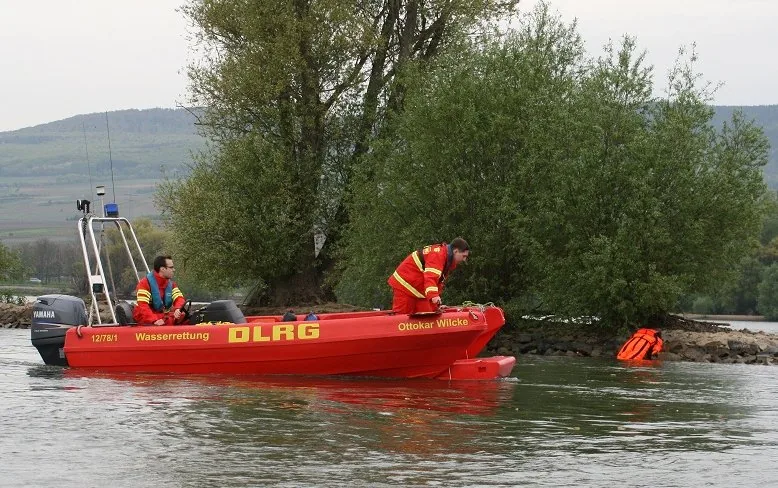 PIONER MULTI III red on the water, seen from the side, with two crew members and rescue equipment near the shore