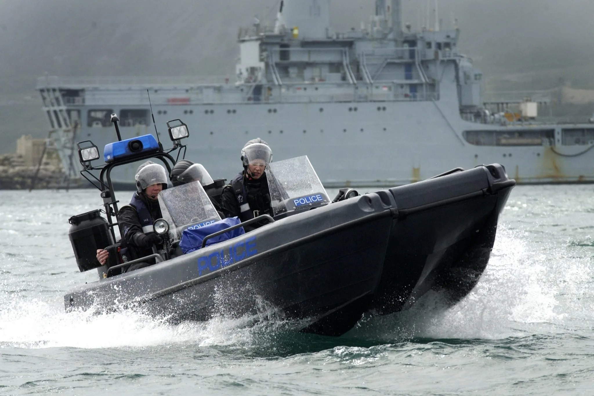 PIONER MULTI III viewed from the bow underway, with two occupants, a central console, and a naval vessel in the background.