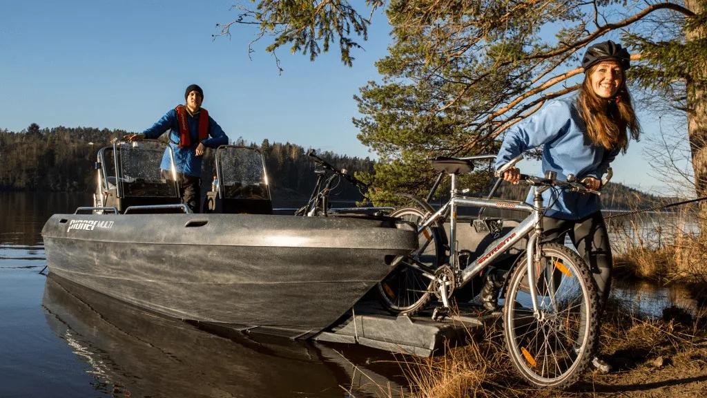 PIONER MULTI III boat in side view next to a lake, with two people and bicycles on board.